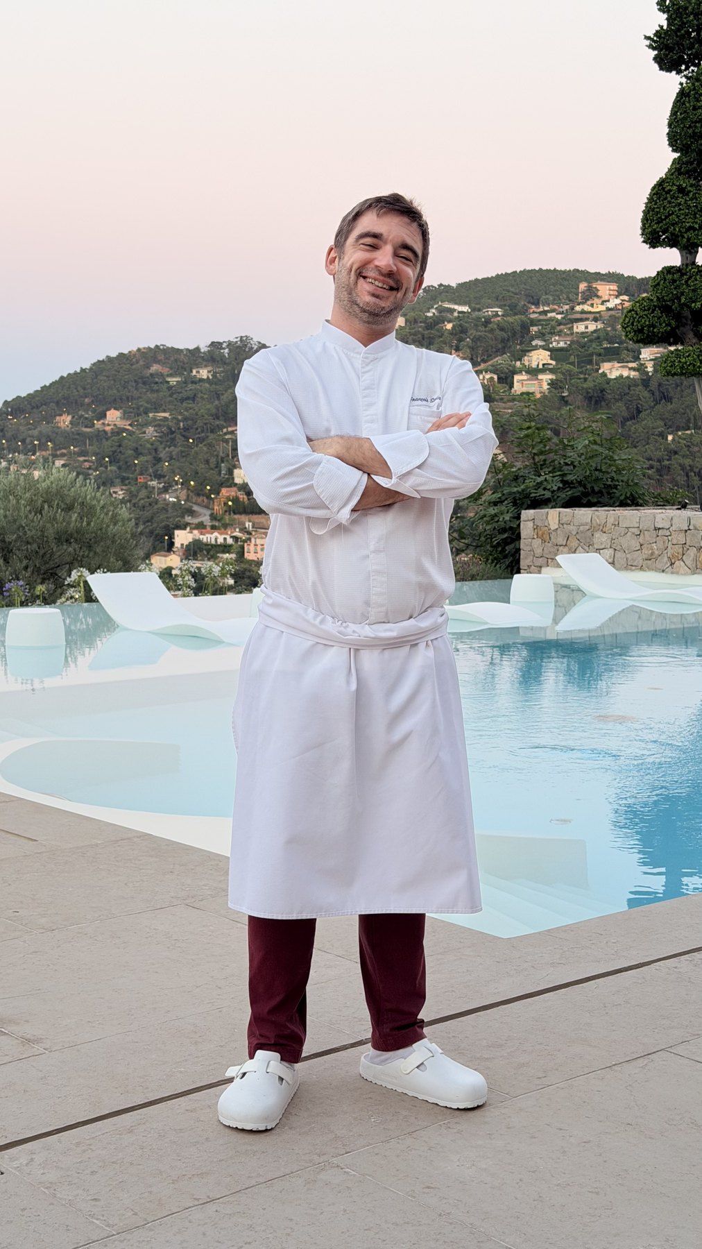 Chef François Savin en veste blanche et tablier, bras croisés au bord d’une piscine à débordement au crépuscule, collines boisées derrière.