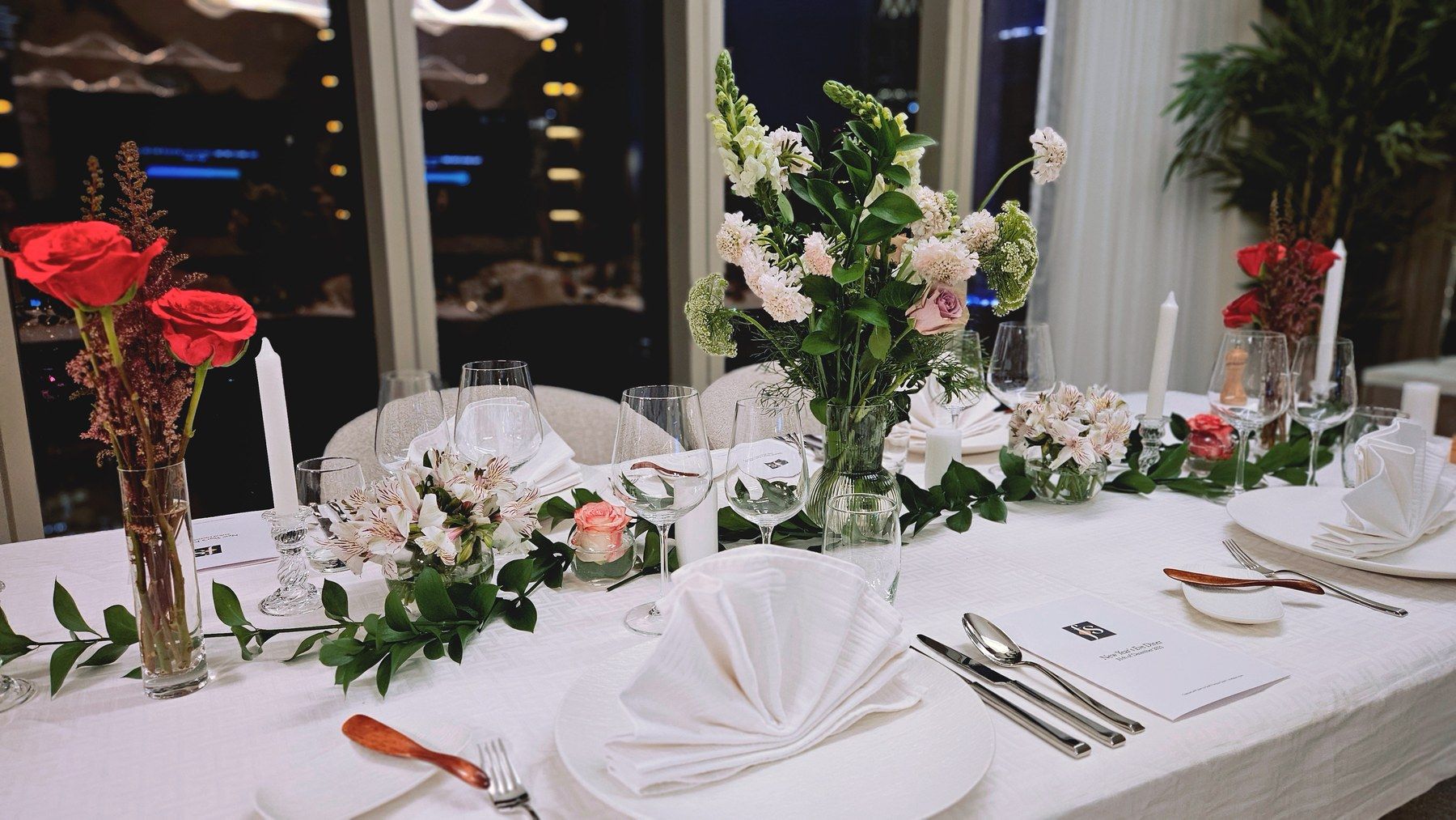 Long white table dressed with roses and candlelight at night.