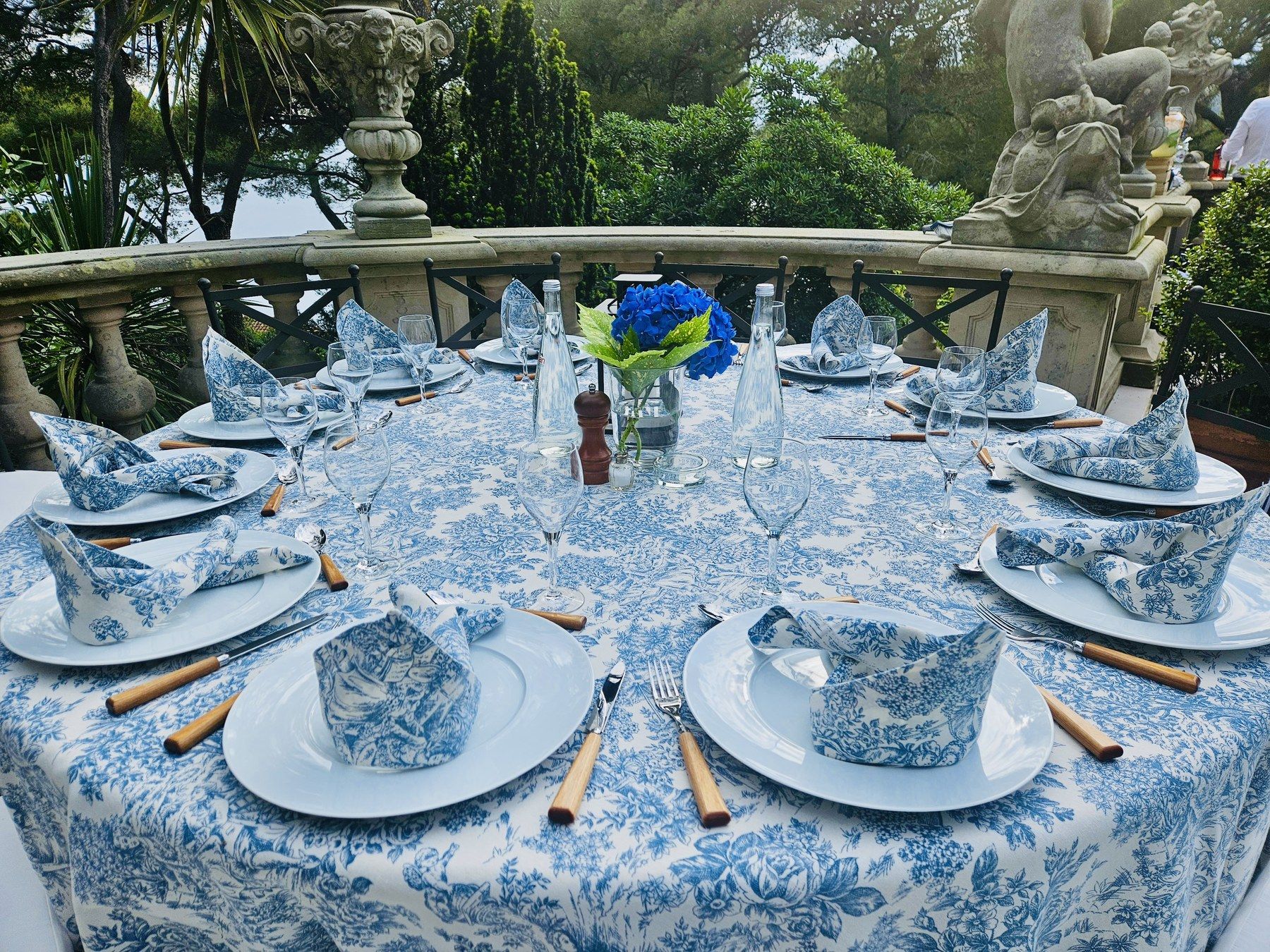 Round table dressed in blue toile de Jouy linen, garden statue behind.